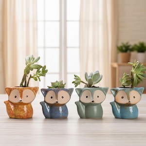 May include: Four ceramic fox-shaped planters, each holding a succulent plant. The planters are in shades of orange, blue, green, and teal, with white and black facial details. The plants have green leaves.