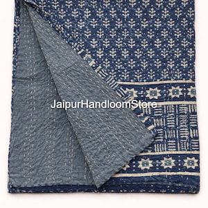 以下が含まれることがあります： 藍色と白の模様の折り畳まれたキルト。キルトは、繰り返しの花の模様と幾何学的な形のボーダーが特徴です。キルトの内側は無地の青色です。「JaipurHandloomStore」の文字が見えます。