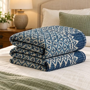 May include: A folded quilt featuring a dark blue and off-white geometric pattern with small floral-like motifs and a zigzag border. The fabric displays visible white stitching throughout, creating a textured, quilted appearance.