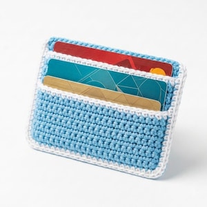 Peut inclure: Un porte-cartes en crochet bleu clair avec une bordure blanche. Cet accessoire rectangulaire comporte deux fentes horizontales contenant chacune une carte en plastique. La carte supérieure est rouge et la carte inférieure est dorée avec un motif géométrique.
