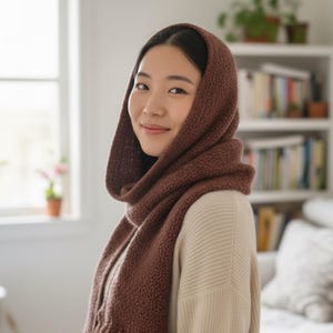 May include: A woman wearing a brown knitted hood and scarf set. The hood is pulled up, framing the face, and the scarf is wrapped around the neck. The knitted texture is visible, and the color is a warm, earthy brown.