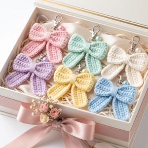 Puede incluir: Un conjunto de seis llaveros de lazo tejidos a ganchillo en rosa pastel, verde menta, crema, lavanda, amarillo y azul claro, cada uno con un cierre de mosquetón de metal plateado y una pequeña perla, dispuestos en una caja de regalo blanca con un lazo rosa.