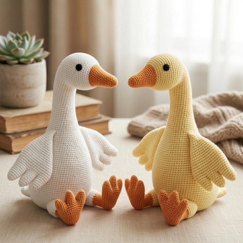 May include: Two handmade crochet geese, one white and one yellow, sit side-by-side. Both have orange beaks and feet, and black eyes. The white goose has a white body, while the yellow goose has a yellow body. The geese are sitting on a light-colored surface.