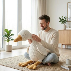 May include: A large, handmade crochet goose plush toy. The goose is primarily white with a yellow beak and feet. It has closed eyes, suggesting a sleeping or relaxed pose. The toy is soft and cuddly, perfect for a child's room or as a decorative item.