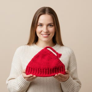 May include: A vibrant red, handmade crochet hat with a pointed top and a tassel. The hat is held in front of a person wearing a white sweater. The hat's texture and color suggest a cozy, winter-themed accessory.