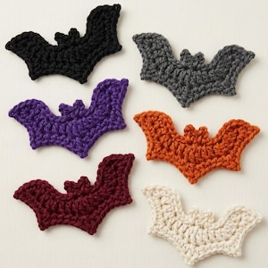 May include: A collection of six crocheted bat appliques in various colors including black, dark gray, purple, orange, deep red, and off-white. Each bat features a distinct wing shape and a small head with pointed ears, crafted with yarn.