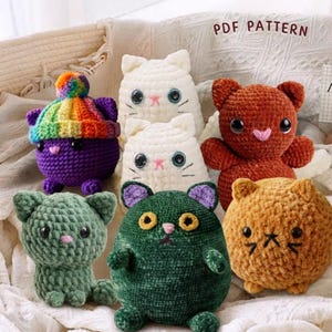 May include: A collection of handmade crochet cat plushies in various colors. The plushies include white, green, orange, and purple cats, some with hats. The text "PDF PATTERN" is visible in the background. These soft toys are perfect for cat lovers.