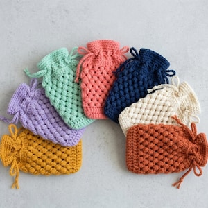 May include: A collection of seven small, rectangular crochet drawstring pouches arranged in a fan shape. Each pouch features a textured bobble stitch pattern and is crafted in a different solid color: yellow, lavender, mint green, coral, navy blue, cream, and rust orange.