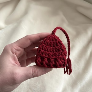 May include: A small, handmade, burgundy crochet tea bag holder. The tea bag holder has a rounded shape with a textured surface and a long, looped handle with a tassel. The item is held in a hand against a soft, neutral background.