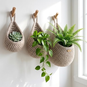 Puede incluir: Tres cestas colgantes de ganchillo beige suspendidas de ganchos de madera en la pared. Cada cesta contiene una planta verde diferente, incluyendo una suculenta, un poto colgante y un helecho. La cesta más grande tiene una etiqueta SET.