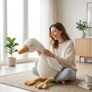 May include: A large, handmade crochet goose in cream and yellow. The goose has a long neck, closed eyes, and detailed feet. The plush toy is sitting on a woven rug in a well-lit room.