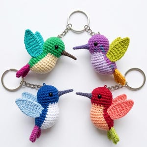 Puede incluir: Cuatro llaveros de colibrí tejidos a ganchillo con hilos de colores vibrantes. Uno es verde y crema, otro morado y lima, otro azul y blanco, y otro rojo y melocotón. Cada ave tiene un pico oscuro, ojos de cuentas negras y una anilla metálica.