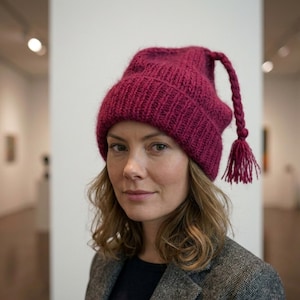 May include: A burgundy knitted beanie hat with a long, braided tassel. The hat is made from a thick, textured yarn and has a ribbed brim. The woman in the image is wearing a grey tweed jacket and a black shirt.