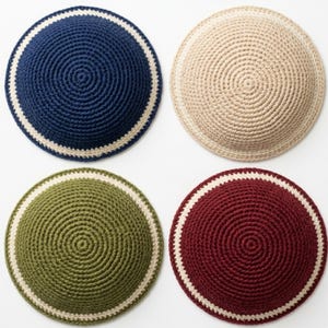 Può includere: Quattro kippah all'uncinetto in blu navy, beige, verde oliva e bordeaux. Ogni kippah presenta un design a spirale e un bordo color crema. I copricapi circolari sono esposti su uno sfondo bianco.