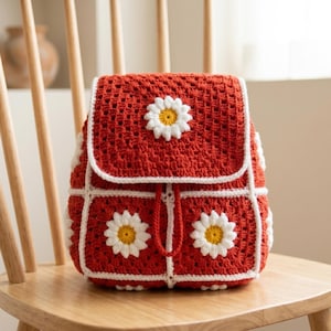 Puede incluir: Una mochila roja de ganchillo hecha a mano con ribetes blancos y detalles de margaritas. La bolsa tiene un cierre de solapa y un cordón. Las margaritas tienen pétalos blancos y centros amarillos. La mochila está sobre una silla de madera.