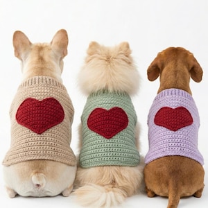 May include: Three dogs wearing handmade crochet sweaters with a large red heart design on the back. The sweaters are in beige, green, and lavender. The dogs are facing away from the camera.