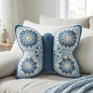 May include: A blue and white crochet butterfly pillow. The pillow is shaped like a butterfly with detailed wings. The wings feature a circular pattern in shades of blue and white. The pillow is on a bed with white pillows.