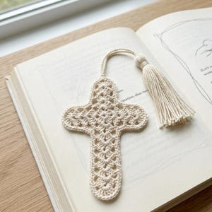 May include: A cream-colored, crocheted cross bookmark with a matching tassel. The bookmark is lying on an open book with visible text. The cross design features an open, lacy pattern. The tassel is attached to the top of the bookmark with a small loop.