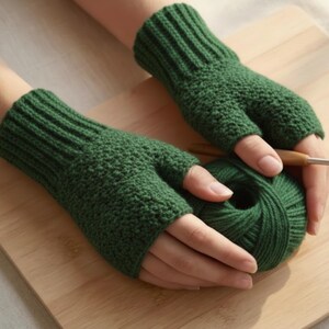 May include: A pair of forest green fingerless gloves made of knitted yarn. The gloves are held over a ball of matching green yarn, with a crochet hook visible. The gloves have a ribbed cuff and a textured stitch pattern.