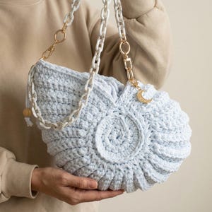 Puede incluir: Un bolso de ganchillo azul claro con forma de concha de nautilo. El bolso cuenta con una correa de cadena de plástico blanco veteado con herrajes de metal dorado y un pequeño dije de media luna de color dorado en el lateral de la abertura.