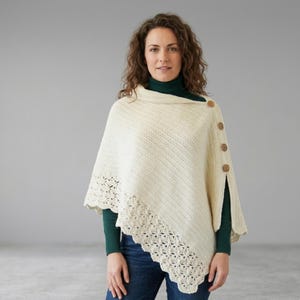 Puede incluir: Poncho de punto color crema con borde festoneado decorativo y botones de madera. El poncho tiene cuello alto y un diseño diagonal. La modelo lleva un cuello alto verde y vaqueros azules.
