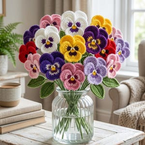Könnte beinhalten: Ein Strauß bunter, gehäkelter Stiefmütterchen in einer durchsichtigen Glasvase. Die Blumen sind in Rot-, Rosa-, Lila-, Gelb- und Weißtönen gehalten, mit grünen Blättern. Die Vase steht auf einem hellen Holztisch.