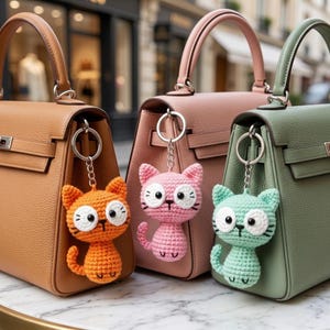 May include: Three structured leather handbags in tan, dusty pink, and sage green, each featuring a silver-toned metal ring attachment. Hanging from each ring is a small, handmade crochet cat keychain in matching orange, pink, and light green yarn.