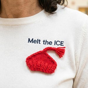 May include: A cream-colored sweater with the phrase "Melt the ICE" in navy blue lettering. A small, red, knitted heart-shaped design with a tassel is attached to the sweater. The sweater is made of a soft, textured material.