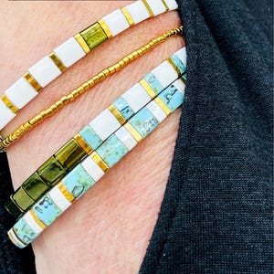 May include: Close-up of several beaded bracelets in white, gold, green, and turquoise. The bracelets are stacked on top of each other. The text "NEW COLLECTION" is at the top of the image.