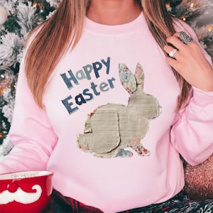May include: A light pink sweatshirt with the words "Happy Easter" in blue lettering. A patchwork bunny design is on the front. The bunny is made of various fabric scraps in neutral tones and floral patterns. The sweatshirt is a crew neck style.
