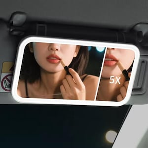 May include: A rectangular car visor mirror with a bright white border and built-in LED lights. The mirror reflects a person applying lipstick. A smaller section of the mirror shows a 5X magnification of the lips.