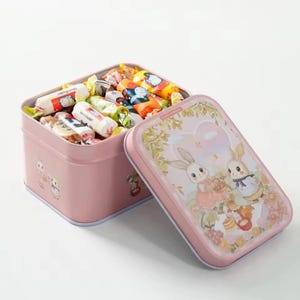 Puede incluir: Una caja de lata rectangular rosa con tapa, llena de caramelos envueltos. La tapa presenta una ilustración de dibujos animados de dos conejos. La caja está abierta, revelando los coloridos caramelos en el interior. La caja es de color rosa claro.