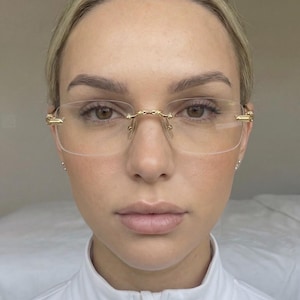 Op de afbeelding: Bril zonder montuur met goudkleurige accenten. De bril heeft een rechthoekige vorm. Het montuur is dun en delicaat, met een brug en veren in goud. De glazen zijn helder. Gedragen door een persoon.