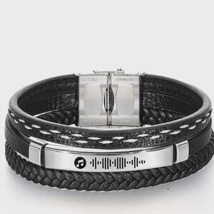 May include: Black leather bracelet with a stainless steel clasp and a silver plate featuring a music note and a sound wave design. The bracelet has multiple layers with braided and stitched details.