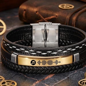 May include: Black leather bracelet with a gold-colored plate featuring a music symbol and a sound wave design. The bracelet has a stainless steel clasp and white stitching details. The bracelet is a multi-layered design.
