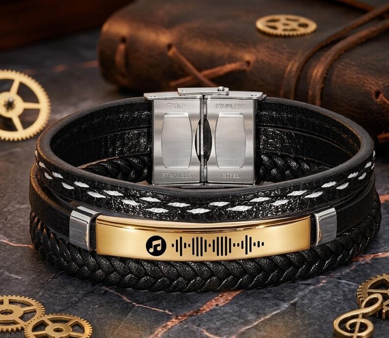 May include: Black leather bracelet with a gold-colored plate featuring a music symbol and a sound wave design. The bracelet has a stainless steel clasp and white stitching details. The bracelet is a multi-layered design.