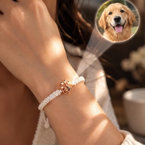 Può includere: Un braccialetto in corda intrecciata bianca con un ciondolo a forma di zampa color oro rosa al centro. Il ciondolo contiene una piccola lente di proiezione circolare che mostra una fotografia di un golden retriever con la lingua di fuori.