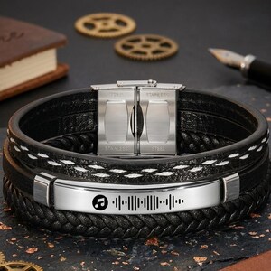 May include: Black leather bracelet with a stainless steel clasp and a silver band featuring a music waveform design. The bracelet has multiple braided layers with white stitching accents. A pen and a book are in the background.