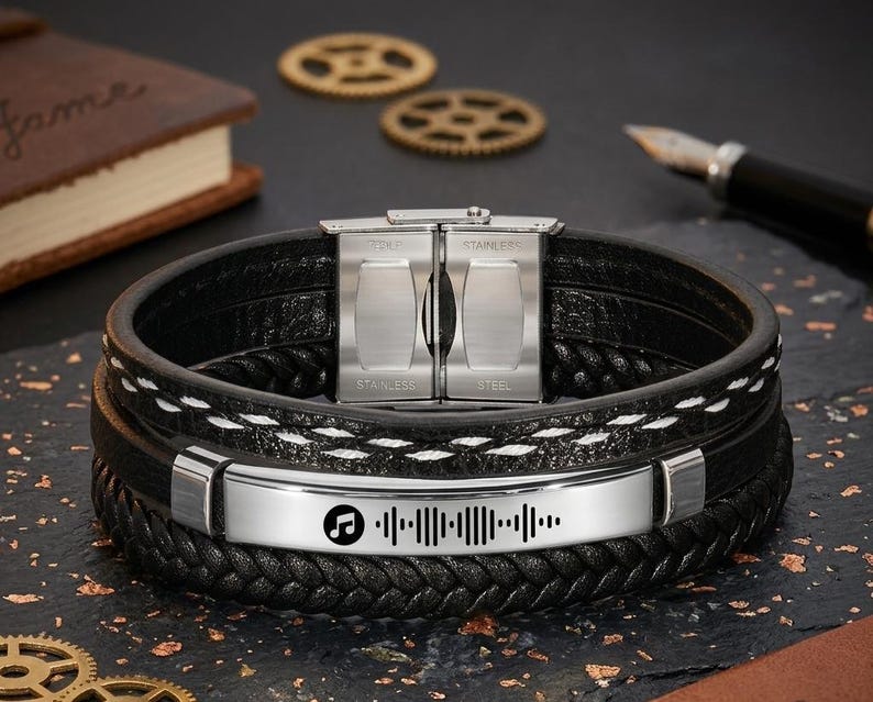 May include: Black leather bracelet with a stainless steel clasp and a silver band featuring a music waveform design. The bracelet has multiple braided layers with white stitching accents. A pen and a book are in the background.