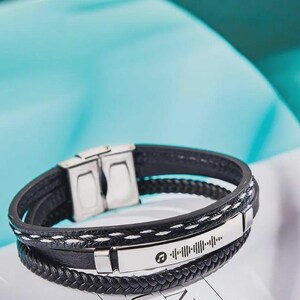 May include: Black leather bracelet with a silver clasp and a silver plate featuring a music note symbol and a barcode-like design. The bracelet has a braided leather band and a stitched leather band.