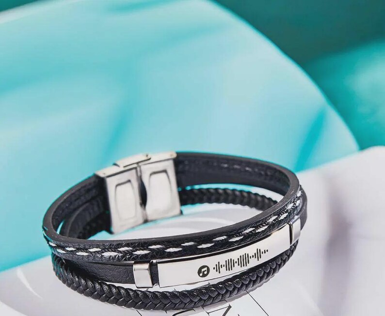 May include: Black leather bracelet with a silver clasp and a silver plate featuring a music note symbol and a barcode-like design. The bracelet has a braided leather band and a stitched leather band.