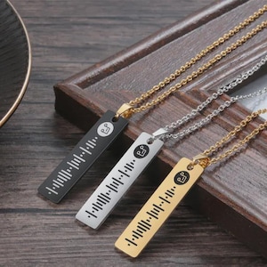 May include: Three pendant necklaces in black, silver, and gold. Each rectangular pendant features a unique barcode design and a musical note symbol. The chains are gold and silver, and the pendants are displayed on a wooden surface.