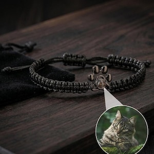 Puede incluir: Pulsera trenzada negra con un dije de huella de pata. El dije tiene una pequeña proyección circular con una imagen de gato. La pulsera se muestra sobre una superficie de madera oscura.
