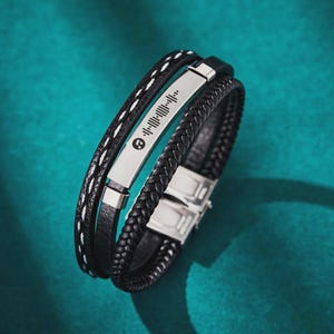 May include: A multi-layered black leather bracelet with a silver metal plate featuring a music code and a music symbol. The bracelet has a braided leather band and a band with white stitching.