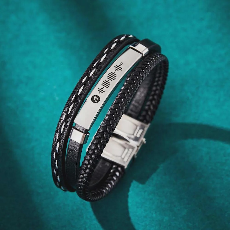 May include: A multi-layered black leather bracelet with a silver metal plate featuring a music code and a music symbol. The bracelet has a braided leather band and a band with white stitching.