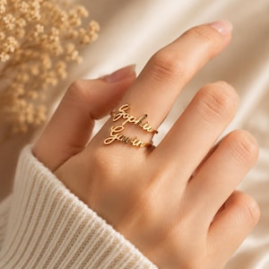 Puede incluir: Un anillo de metal dorado con dos placas de nombre superpuestas en letra cursiva. La placa superior muestra el nombre Sophia y la placa inferior muestra el nombre Gavin. El anillo tiene una banda delgada y pulida.