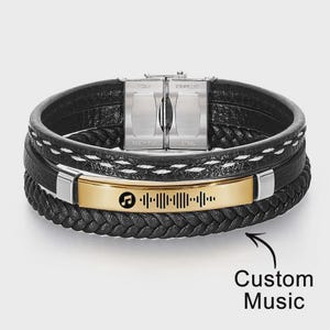 May include: Black leather bracelet with a gold-colored plate featuring a music-themed design. The bracelet has multiple layers and a stainless steel clasp. The text "Custom Music" is in the image.