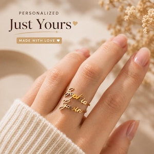 May include: A gold-toned ring featuring two stacked, cursive-style name plates displaying the names Sophia and Gavin. The ring is worn on a finger, and the image includes the text Personalized Just Yours and Made With Love.