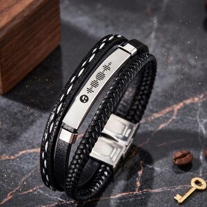 May include: Black leather bracelet with a silver metal plate featuring a Spotify code. The bracelet has multiple strands, including a braided section and one with white stitching. A metal clasp secures the bracelet.