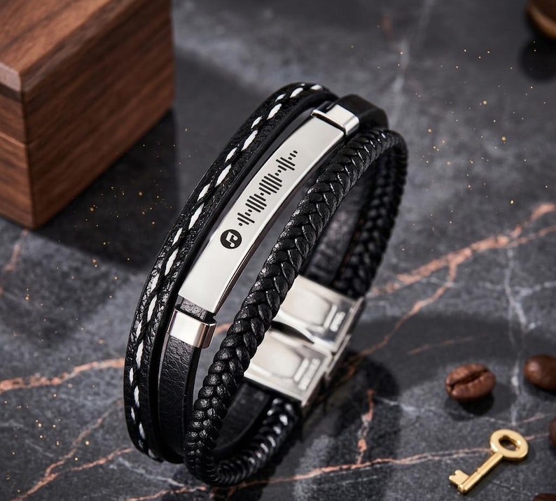 May include: Black leather bracelet with a silver metal plate featuring a Spotify code. The bracelet has multiple strands, including a braided section and one with white stitching. A metal clasp secures the bracelet.